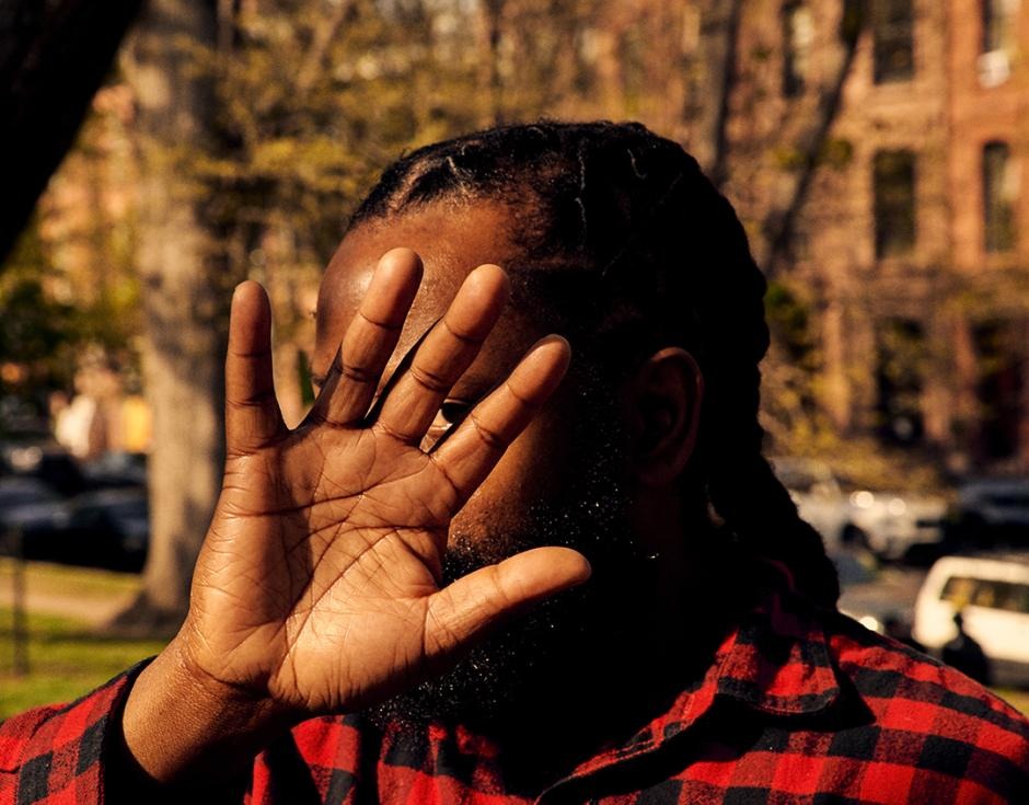 A photograph of billy woods. He wears a red flannel and conceals his face with his hand.